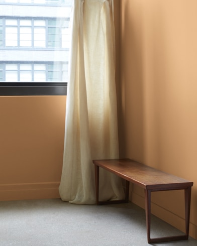 In a Honeycomb-painted room with a wooden bench, a gauzy white curtain hangs next to an open window.